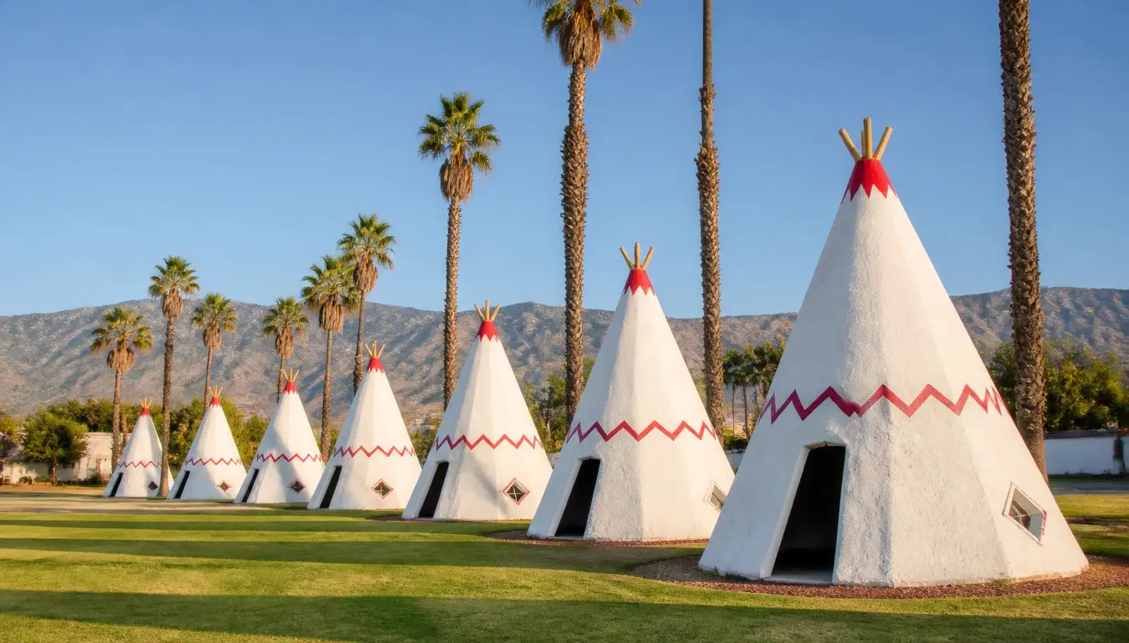 Row of white teepee-shaped roadside cottages and tall palms along historic Route 66 in Rialto, CA with the mountains behind
