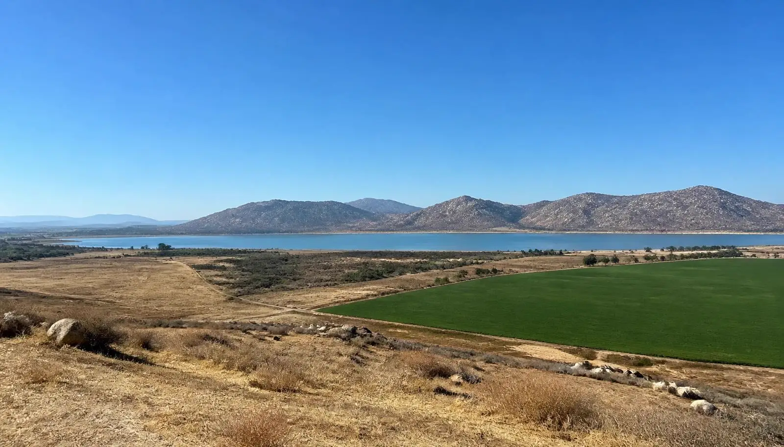Flat Perris, CA valley with dry grassland, a green irrigated field, and the reservoir below rocky hills