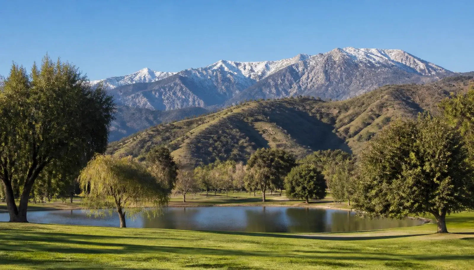 Park lake and green lawn in Yucaipa, CA with oak-covered foothills and the snow-topped ridge behind